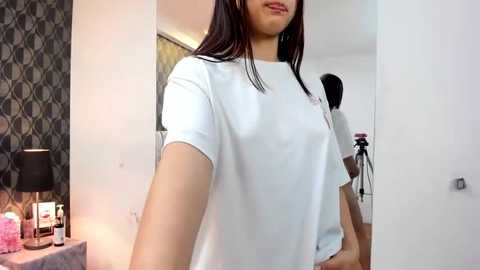 Media: A video of a young Asian woman with long black hair, wearing a white T-shirt, standing indoors, with a black lamp, patterned wallpaper, and a mirror in the background.