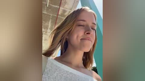 Media: A video of a fair-skinned young woman with long blonde hair, wearing a white, off-the-shoulder knit top, smiling, set against a bright, sunlit, modern architectural backdrop.