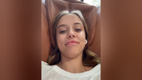 Media: Video of a young Caucasian woman with light brown hair, lying on a brown leather recliner, wearing a white knit sweater. She has a relaxed, content expression.