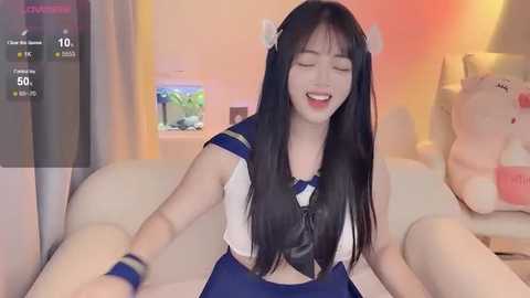 Media: Video of an Asian woman with long black hair in a sailor outfit, smiling, sitting on a couch with a stuffed animal, in a cozy, warmly lit room.