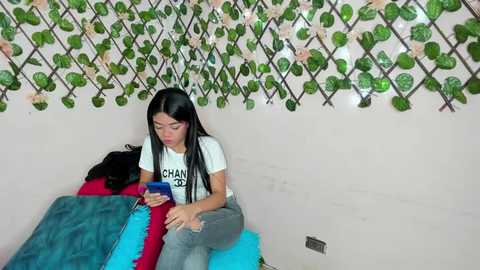 Media: A video of a young woman with long black hair, wearing a white T-shirt and ripped jeans, sitting on a turquoise bed, holding a blue phone, surrounded by a green leafy wall decor.