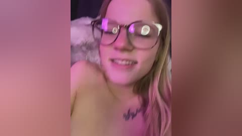 Media: A blurry, topless video of a young woman with fair skin and long blonde hair, wearing glasses, smiling, and showing a tattoo on her chest.