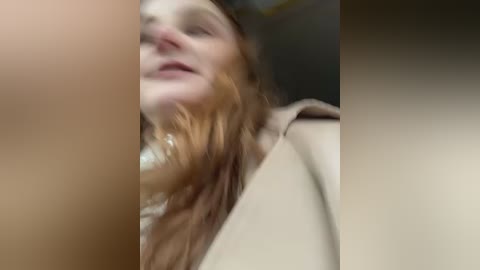 Media: A blurred video of a young woman with long, wavy auburn hair, wearing a beige coat, seen from a low angle, with a partially obscured face and blurred background.