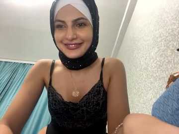 Media: Video of a smiling woman with light brown skin, wearing a black lace camisole and black hijab with a white scarf underneath, in a room with teal curtains and textured wallpaper.