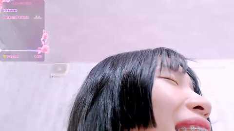 Media: Video of a young Asian woman with straight black hair, closed eyes, and slightly open mouth, taken from a low angle, with a blurred pink and white background.