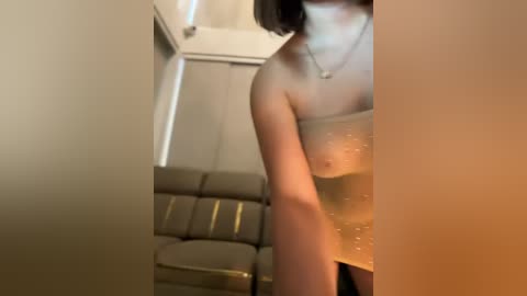 Media: A blurred video of a partially nude woman with medium skin tone, wearing a glittery dress, standing in a beige, minimalist room with a couch and white walls.