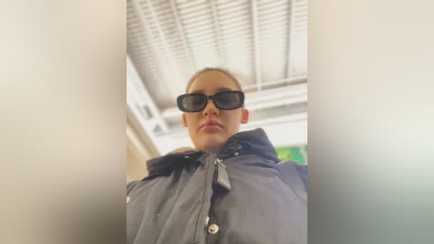 Media: Video of a woman with light skin, short hair, and black sunglasses, wearing a dark jacket, taken from a low angle in a bright, industrial setting with a white ceiling and fluorescent lights.