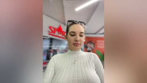 Media: Video of a Caucasian woman with light skin, dark hair tied back, wearing a white ribbed turtleneck sweater, standing in a supermarket. Background shows red and white signage, shelves, and blurred products.