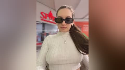 Media: A video of a young woman with fair skin and long brown hair in a ponytail, wearing a white ribbed turtleneck sweater and black sunglasses. She stands in a brightly lit indoor setting with blurred background.