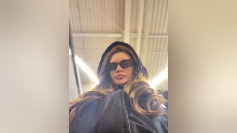 Media: A video of a woman with long, wavy brown hair, wearing black sunglasses and a dark coat, standing in a modern, industrial building with high ceilings and white beams.