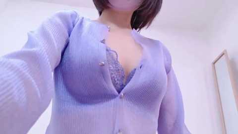 Media: A video of a light-skinned woman with short, dark hair, wearing a sheer, purple button-down blouse that reveals a lacy bra underneath. The background is a minimalist, white-walled room with a wooden door.