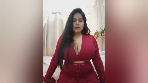 Media: Video of a curvy Latina woman with long black hair, wearing a red V-neck jumpsuit, sitting on a bed with white sheets and beige curtains in the background.