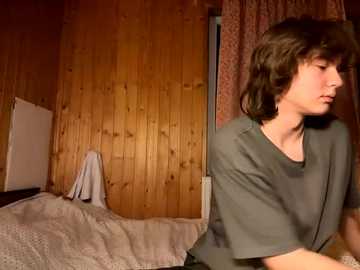 Media: Video of a young woman with shoulder-length brown hair, wearing a green T-shirt, sitting on a bed with a white blanket in a rustic wooden-paneled room with patterned curtains.