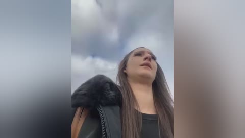 Media: Video of a woman with long brown hair and light skin, wearing a black leather jacket with a fur collar, gazing upward at a cloudy sky.