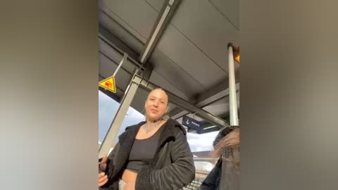 Media: Video of a blonde woman with a beard, wearing a black tank top and jacket, standing in a subway train, holding a pole.