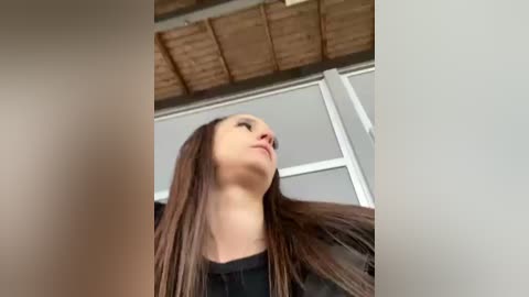 Media: Video of a young woman with long, straight, dark brown hair, wearing a black top, looking upward, against a wooden ceiling and white-framed windows.