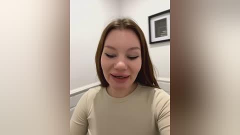 Media: Video of a young woman with fair skin, long brown hair, and light makeup, wearing a beige long-sleeve top, standing in a hallway with white walls and framed artwork.