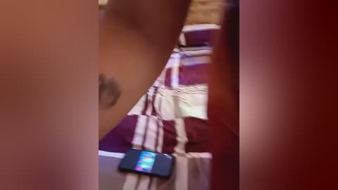 Media: A blurry, close-up video of a person's upper body, featuring a tattoo on the right side, holding a smartphone with a colorful screen. The background shows a striped bedspread in red and white.