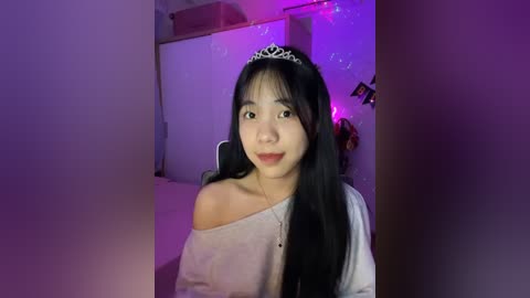 Media: Video of an East Asian woman with long black hair, wearing a silver tiara, off-shoulder gray top, and pink lipstick, in a dimly lit room with purple lighting and fairy lights.