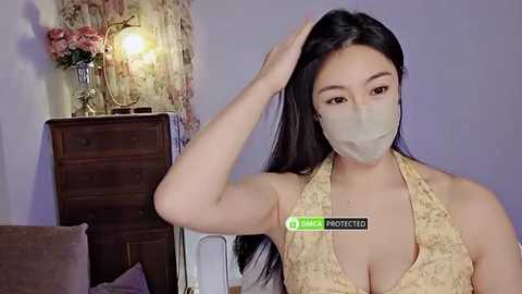 Media: A video of an East Asian woman with long black hair, wearing a yellow halter top, beige face mask, and a green \"Vera Protection\" label. She sits in a dimly lit room with a wooden dresser and flowers.