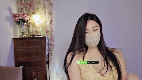 Media: Video of a young Asian woman with long black hair, wearing a beige face mask and a sleeveless lace top, standing in a cozy room with a floral-patterned curtain, a wooden dresser, and a lit lamp.