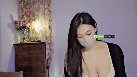 Media: A video of an Asian woman with long black hair, wearing a face mask, a black cardigan, and a revealing white lace bra, standing in a dimly lit room with floral wallpaper and a wooden dresser.