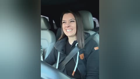 Media: Video of a smiling woman with straight, dark brown hair, wearing a black jacket and seatbelt, seated in a car with gray interior.