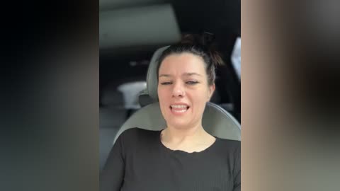 Media: Video of a smiling woman with dark hair in a bun, wearing a black shirt, seated in a car with blurry background.