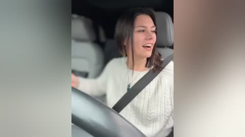 Media: A video of a young woman with shoulder-length brown hair, wearing a white cable-knit sweater, smiling while driving a car with a seatbelt on. The background is blurred, emphasizing the driver's focused expression.