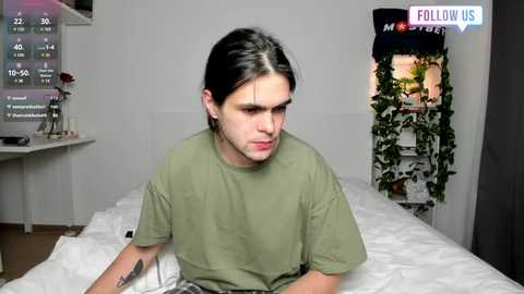 Media: A video of a young man with shoulder-length black hair, wearing a green t-shirt, sitting on a bed in a minimalistic, modern bedroom. The room has a large screen displaying a weather forecast and a green ivy plant in the background.