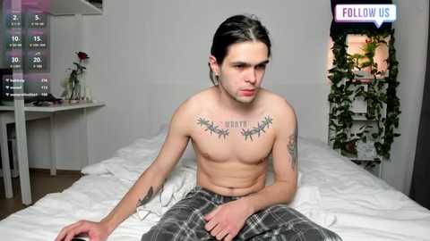 Media: Video of a shirtless man with long black hair, tattoos, and plaid pajama pants, sitting on a bed in a minimalist bedroom with a digital timer and a green plant.