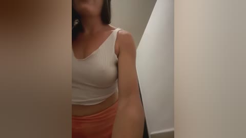 Media: Video of a woman with medium skin tone and long dark hair, wearing a white ribbed crop top and orange pants, standing in a mirror-lit dressing room.