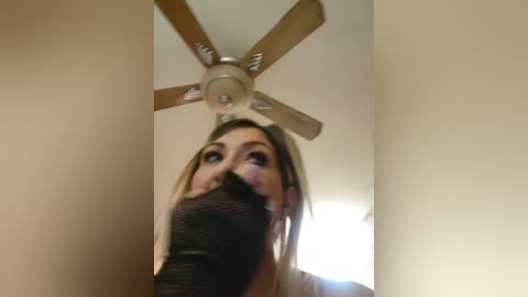 Media: Video of a young woman with blonde hair, wearing a black cloth over her mouth, standing indoors near a ceiling fan.