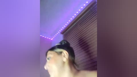 Media: Video of a woman with a high bun, partially obscured, in a room with purple LED strip lights and a closed window with blinds.