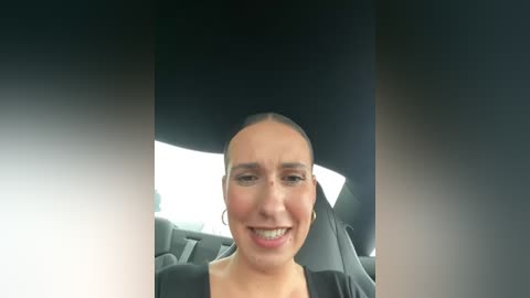 Media: Video of a smiling woman with short black hair, wearing a black top, sitting in a car with black seats. The background is blurred and dark.