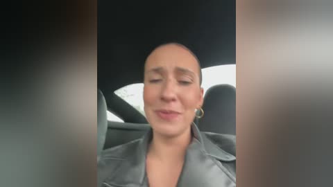 Media: A video of a bald, light-skinned woman with closed eyes, wearing a black leather jacket, seated in a car. The background shows the car's interior.