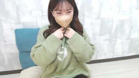 Media: Video of a young Asian woman with long dark hair, wearing a beige mask and a green sweater, sitting indoors against a white, patterned background with a blue cushion.