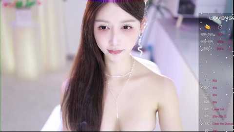 Media: A video of an East Asian woman with long, straight dark hair and fair skin, wearing a purple headband, a pearl necklace, and a white top. She has a slender build and is smiling softly. The background is a softly blurred, indoor setting.