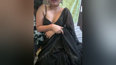 Media: Video of a plus-sized South Asian woman in a black, sheer, low-cut sari, seated on a bed with a patterned green sheet. Her hands are clasped under the sari's folds.