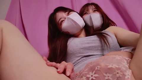 Media: Video of two Asian women with long hair, wearing white surgical masks, lying together on a pink sheet. One wears a light gray tank top, the other a pink lace bra. They have fair skin.