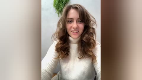 Media: Video of a young Caucasian woman with wavy, shoulder-length brown hair, smiling, wearing a cream-colored, ribbed turtleneck sweater, against a light gray wall with a green plant hanging above.