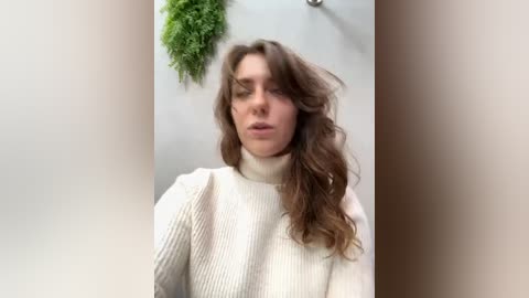 Media: A video of a young woman with long, wavy brown hair, wearing a white ribbed sweater, against a light gray wall with a green potted plant.