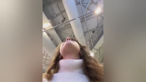 Media: Video of a young woman with long, wavy brown hair, wearing a white ribbed sweater, gazing upward with a serene expression, surrounded by industrial ceiling pipes and lighting in a modern, open space.