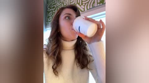 Media: Video of a young woman with wavy brown hair, drinking from a white coffee mug, wearing a cream-colored turtleneck sweater, indoors with a green, white-patterned tapestry in the background.