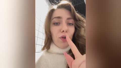 Media: Video of a young woman with light skin and brown hair, wearing a white turtleneck sweater, with a finger to her lips, taken in a tiled bathroom.