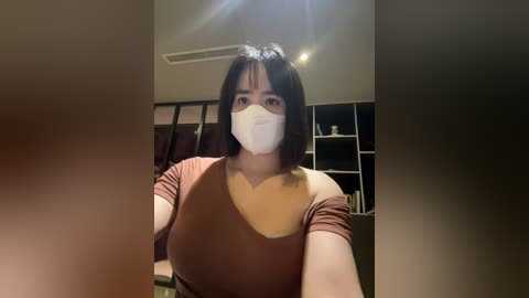 Media: Video of an Asian woman with medium-length black hair and fair skin, wearing a white face mask, brown off-shoulder top, and sitting indoors in a dimly lit room with a bookshelf in the background.