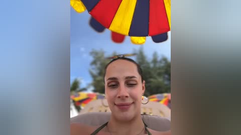 Media: Video of a smiling woman with dark hair, wearing a green bikini, under a colorful striped beach umbrella. Background shows beach chairs and a clear blue sky.