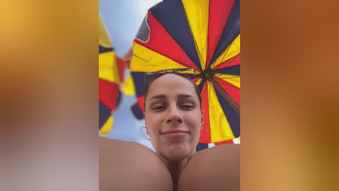 Media: Video of a woman with light skin and brown hair, smiling under a large, colorful umbrella with red, yellow, and black stripes, against a blurred background.