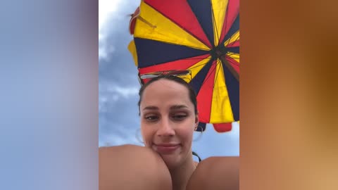 Media: Video of a smiling woman with light brown skin, wearing a black bikini top, holding a large, colorful umbrella with yellow, red, and black stripes, set against a cloudy sky background.