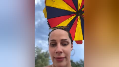 Media: Video of a woman with light skin, dark hair pulled back, wearing sunglasses, holding a vibrant yellow, red, and black umbrella against a cloudy sky.